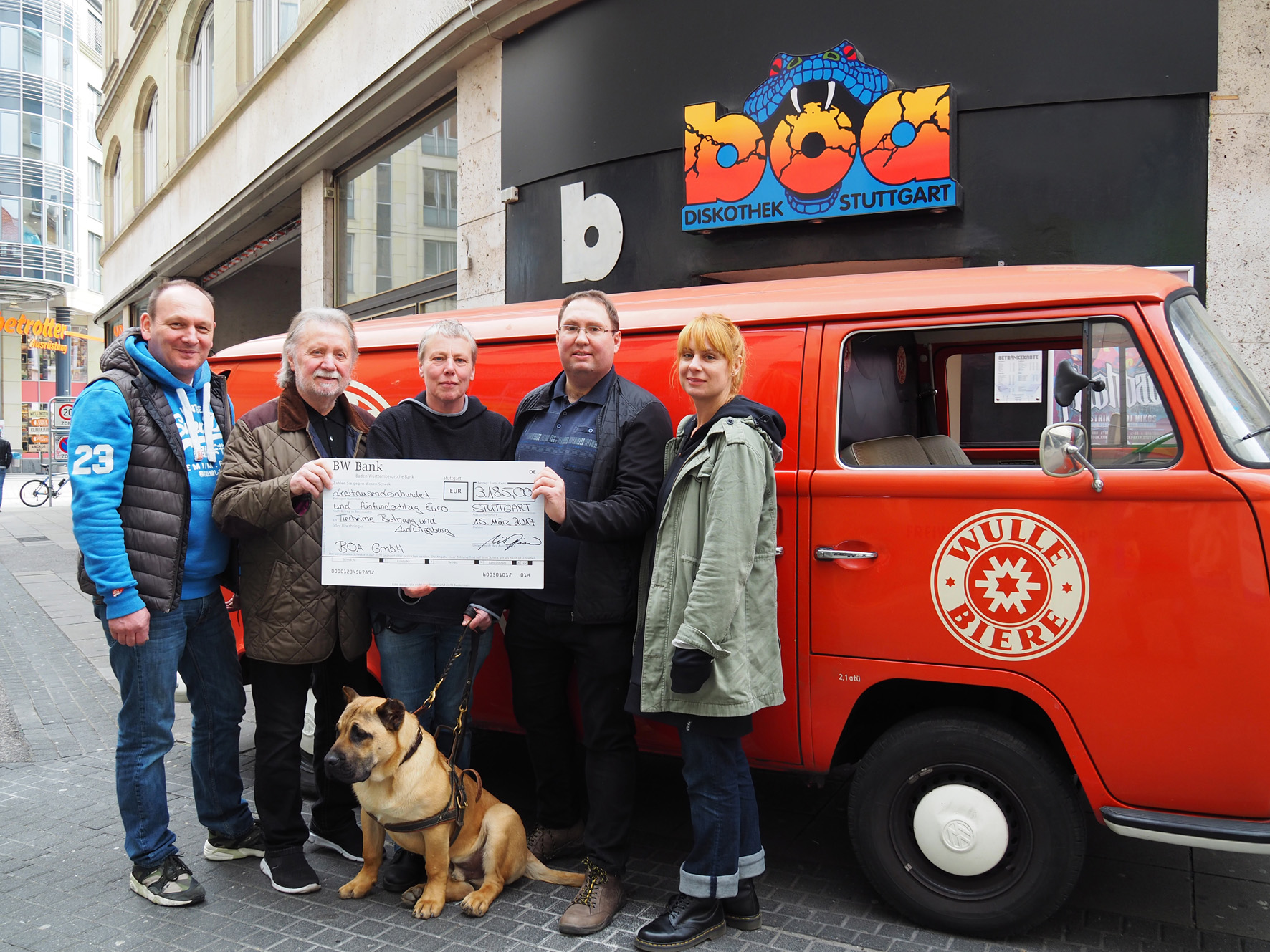 Fünf Personen und ein Hund stehen vor einem roten Lieferwagen und halten einen großen Spendenscheck hoch.