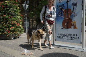 Eine Frau führt einen Schaeferhund an einem Futterbeutel vorbei. Im Hintergrund ist ein Plakat